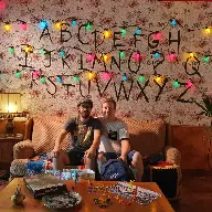 Two beautiful guys sitting in a couch which is on the set of Stranger Things with Christmas alphabet lights